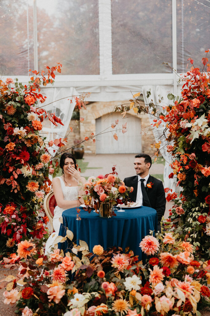 Autumn wedding reception at Pittsburgh Botanic Garden under a clear-top tent with midnight blue velvet linens, wood cross-back chairs, and lush floral arrangements featuring local dahlias and fall foliage.